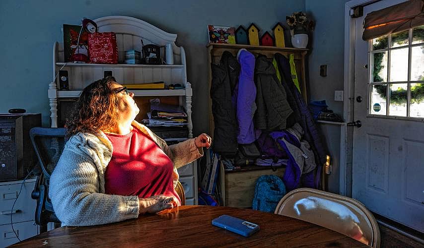 A woman sitting at a kitchen table, looking toward a sunlit door with coats hanging nearby
