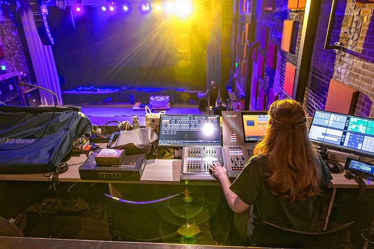 A woman managing theater lighting controls in a booth, facing a brightly lit stage.