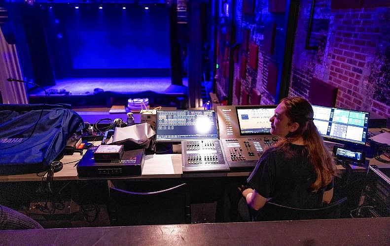 A woman at a control desk in a dimly lit theater, facing multiple screens and equipment.