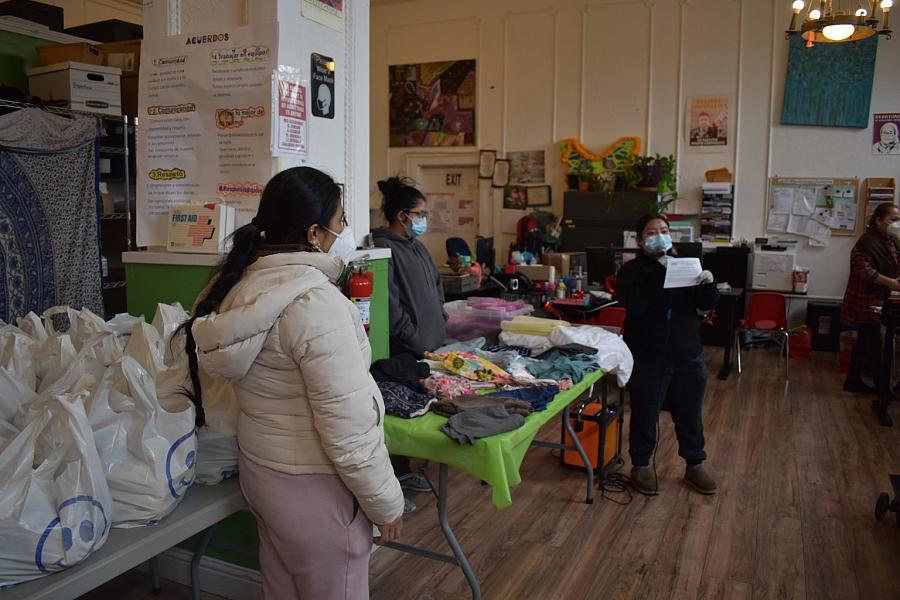 Women wearing masks gather around tables with groceries and clothes inside a community center.