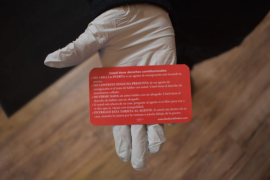 A gloved hand holds a red card listing constitutional rights for immigrants in Spanish.