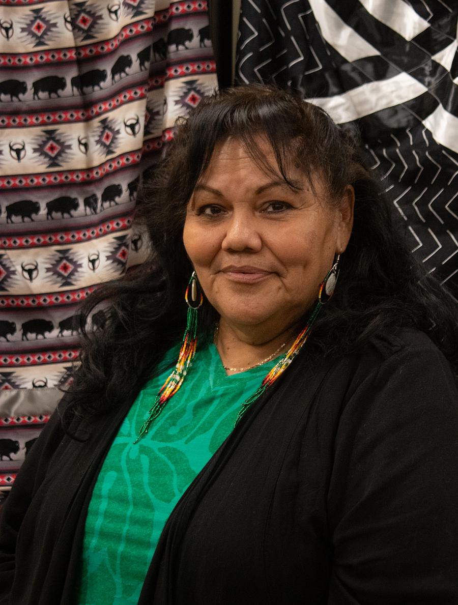 A Native woman with long dark brown hair smiles at the camera in front of draped tapestries