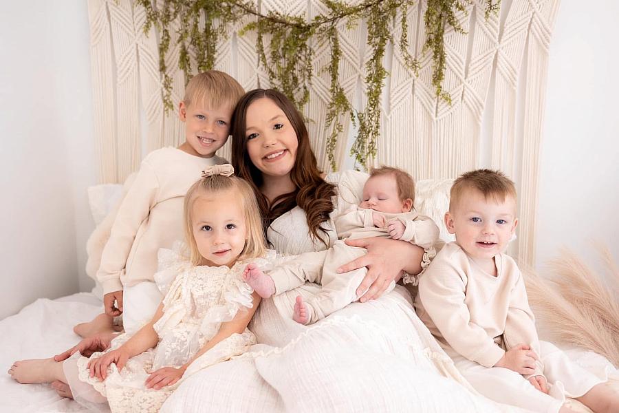 A woman sits on a bed smiling, surrounded by four young children, including a baby in her arms, all dressed in light-colored clothes with soft, natural decor in the background.