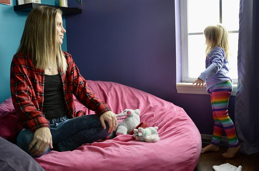 A woman sits on a pink bean bag while a young girl in rainbow leggings looks out a window in a brightly colored room.