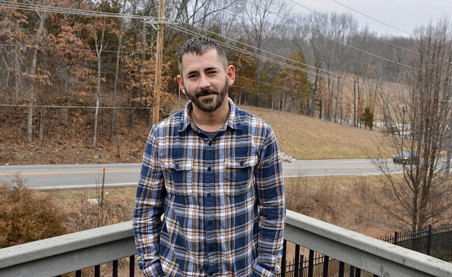 A man in a plaid shirt stands on a balcony, with a rural road and leafless trees in the background on an overcast day.