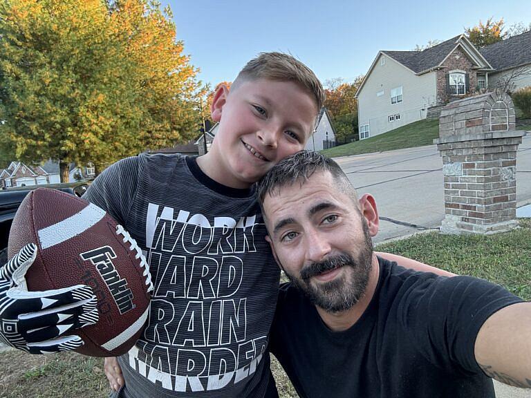 A man and a smiling boy pose together outdoors, with the boy holding a football and wearing a shirt that says “Work Hard Train Harder"