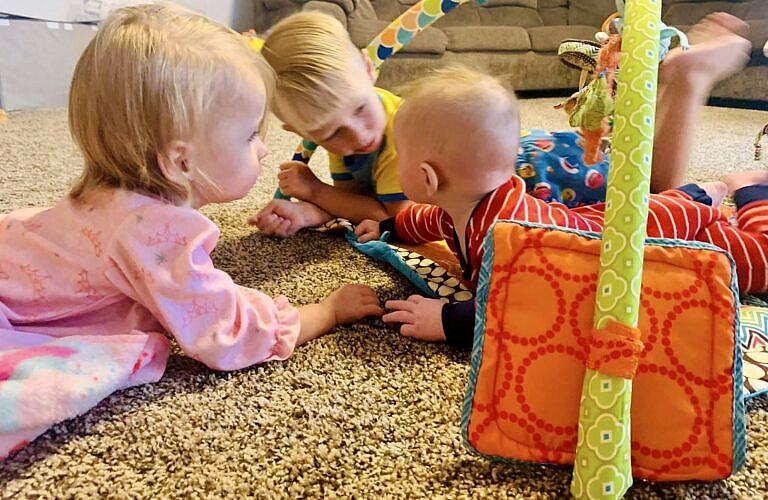 Three young children lie on a carpeted floor, playing and interacting near a colorful baby activity gym.