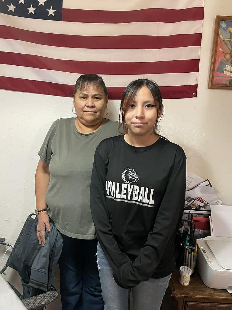 Two women, one older and one younger, pose indoors in front of an American flag.