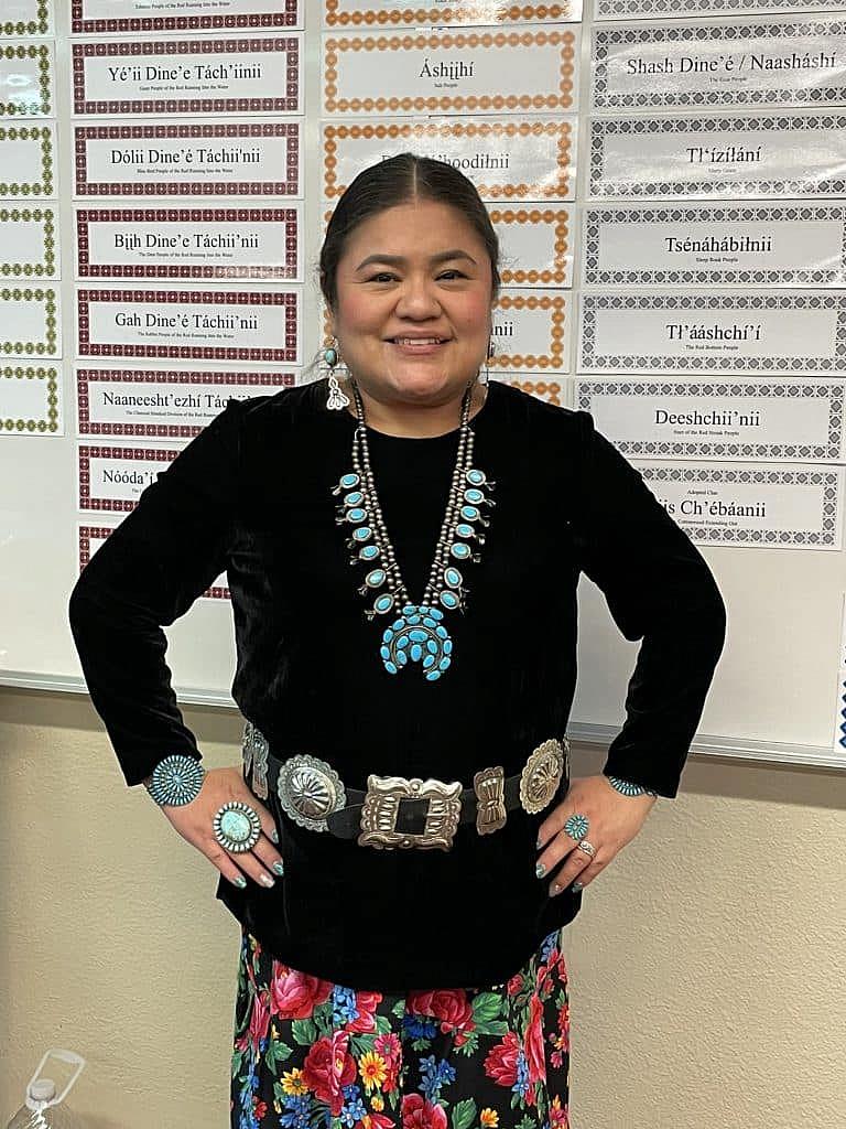 Woman wearing traditional jewelry and floral skirt smiles in front of a classroom wall with Navajo clan names.