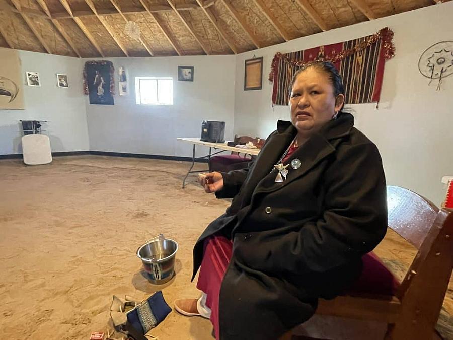 Woman in traditional attire sits on a bench inside a round room with sand floor and cultural decorations.