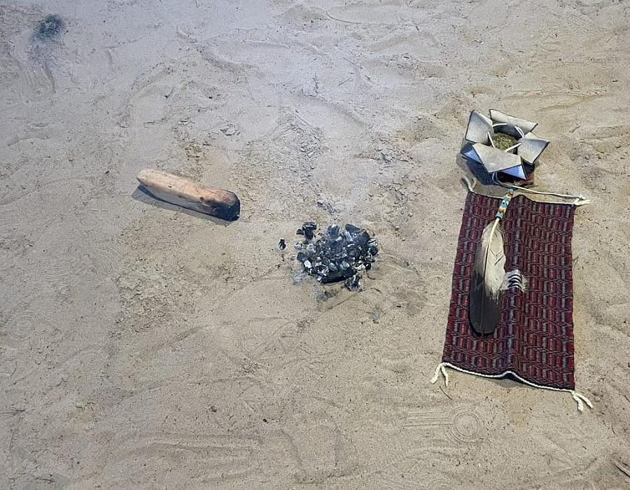 Burned log, ashes, feather, and ceremonial items arranged on sand for a cultural practice.
