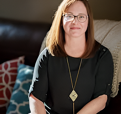 A woman with straight brown hair and glasses sits on a couch, wearing a black top and a long pendant necklace.