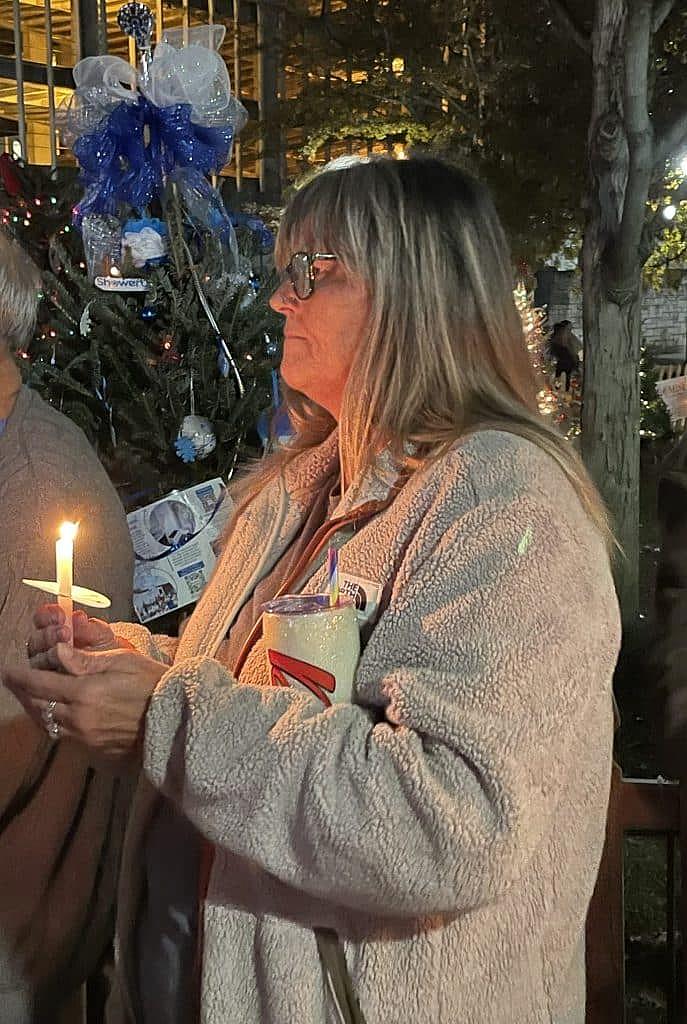 Woman holds candle during evening vigil, with holiday lights and tree behind her.