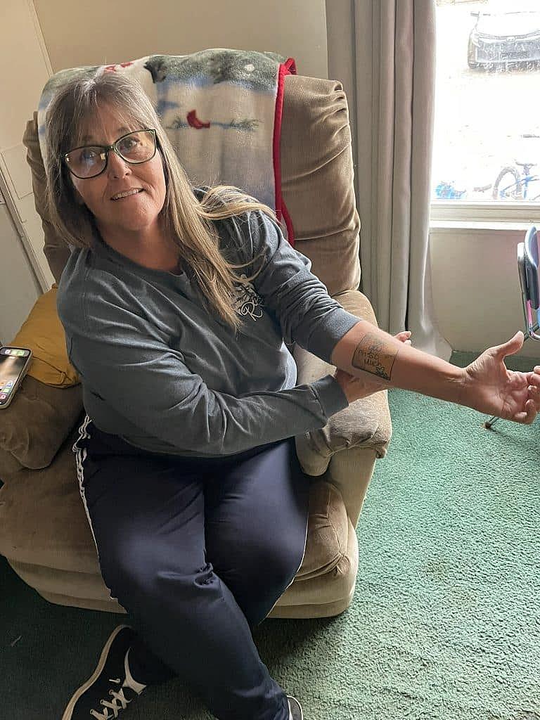 A woman sitting in a recliner at home shows a tattoo on her forearm while smiling; she wears glasses and a long-sleeve shirt.