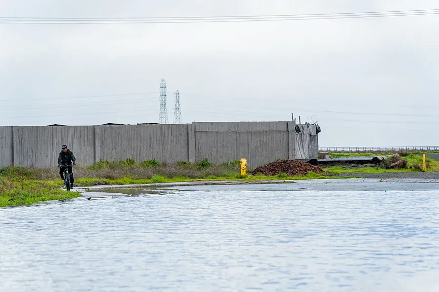 Cyclist rides past a fenced industrial site near a flooded path.