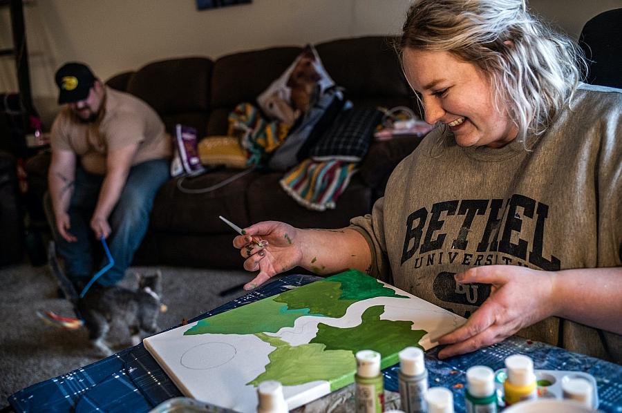 A woman smiling looking at her canvas and her partner in background sitting on couch.