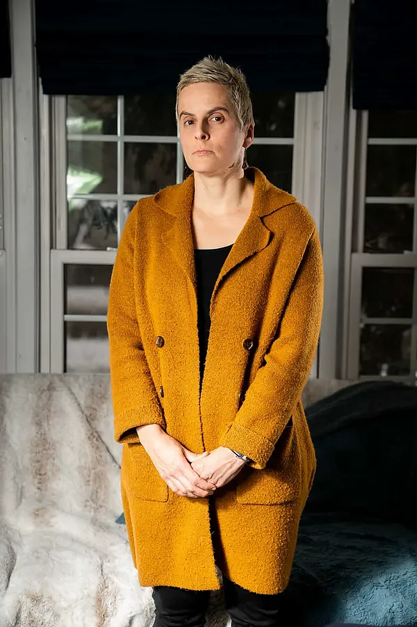 A woman with short, light hair stands indoors in front of large windows, wearing a mustard-yellow coat over black clothing. She looks serious and composed, with hands clasped in front.