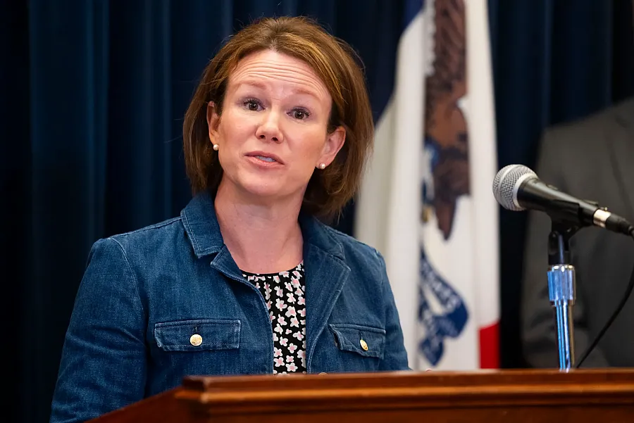 Woman in a denim jacket speaks at a podium with a microphone, standing in front of a dark curtain and a partially visible state flag.
