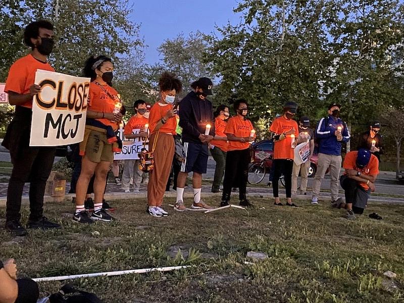 Activists in a park wearing orange clothes; one of them holding board saying "CLOSE MCJ" protesting for closure of MCJ.