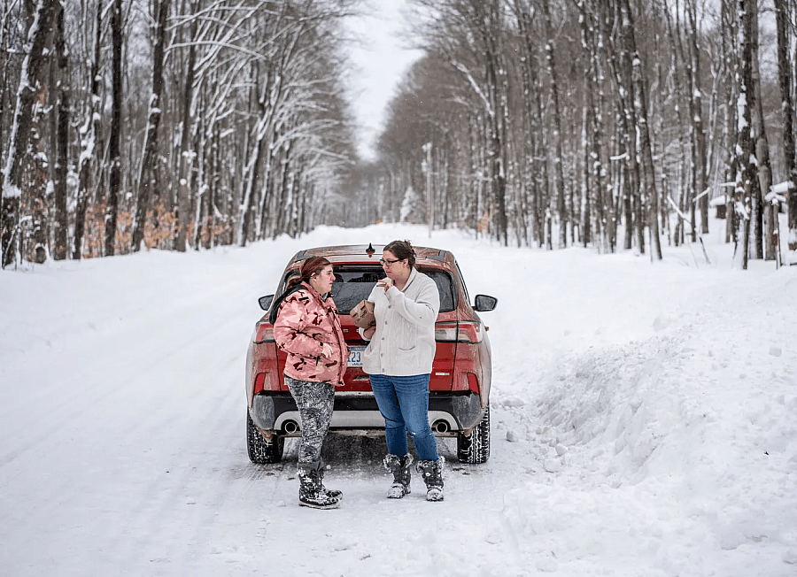 Two people standing behind a red car on a snowclad road with deciduous tree on either sides of the road