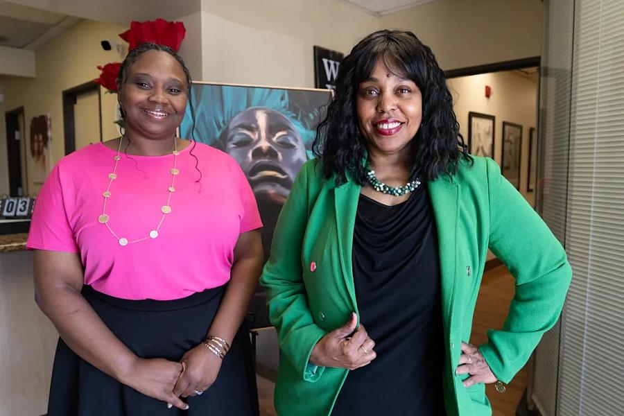 Two women posing indoors, one in a green blazer and one in a pink top.