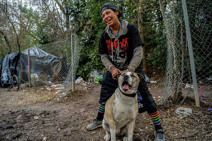 Smiling person holds a dog in an outdoor encampment area near a fence and tents.