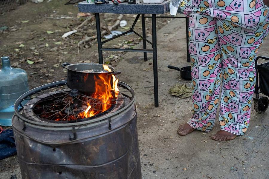 Close-up of a pot over flames on a makeshift barrel stove with woman’s feet nearby.