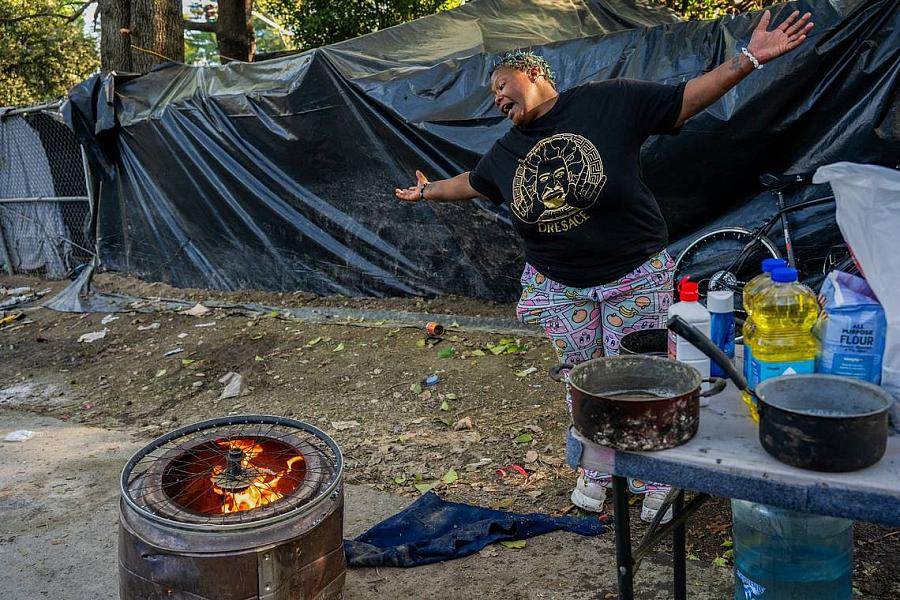 Woman gestures animatedly near a barrel stove and cooking supplies at a campsite.