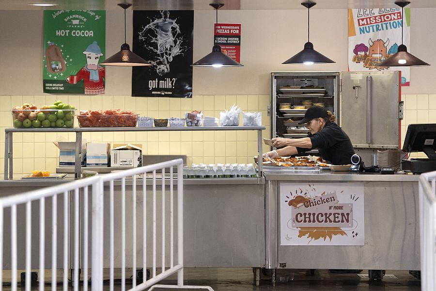 A cafeteria worker serves lunch behind a counter lined with fruit and milk cartons.