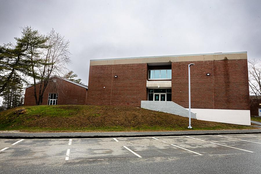 A red brick school building with minimal windows and a small staircase leading to double doors, seen from an empty parking lot on a cloudy day.