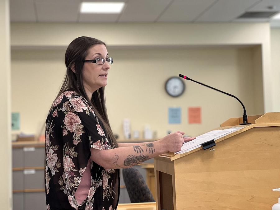 A woman with long hair and glasses speaks at a podium with a microphone, holding papers, in what appears to be a public meeting or hearing room.