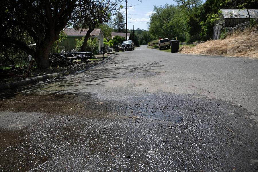 A small puddle of water spreads across a residential street, likely from runoff or a leak, with trees, houses, and parked vehicles visible in the background under a sunny sky.