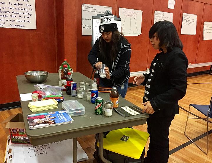 Two volunteers scoop sugar into jars to see how much is included in popular beverages.