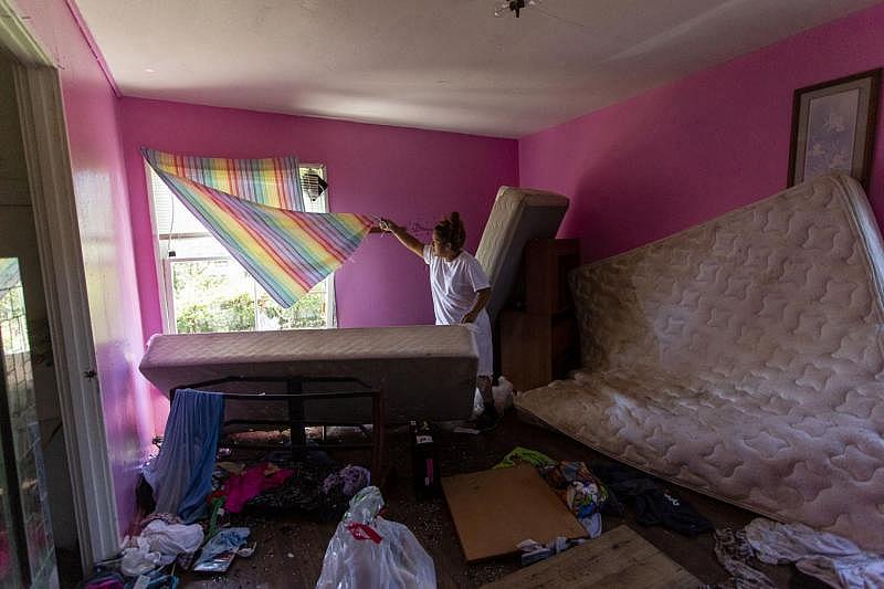 Barrientes lifts a curtain to show a broken window. The mother said she attempted to barricade the window with a mattress during Hurricane Harvey, but the winds were too strong. Broken glass, clothing and a water-damaged mattress are scattered across the bedroom floor.
