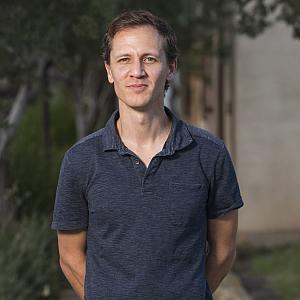 A man stands outdoors in front of trees and a building, wearing a short-sleeved, dark blue polo shirt. He has short brown hair and a neutral expression, with his hands behind his back.