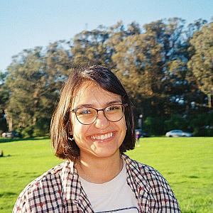 A young women with a brown bob and glasses stands smiling in a field and a plaid shirt.