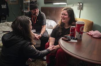 A woman smiles while speaking with two others in a cozy room. One person holds her hands supportively. A soda, coffee cup, and keys rest on the nearby table.
