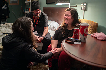 Three people sitting inside a room, one besides a table, one on a bed talking to each other.