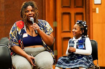 A child seated next to person speaking into a microphone