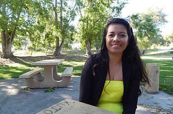 A young woman with long dark hair sits at a picnic bench smiling in a park