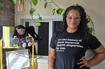 A young Black woman in glasses stands hand on hip inside a doula clinic