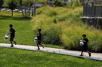 Three children run through a park with bags in their hands.