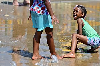 A child plays with a friend in a sprinkler.