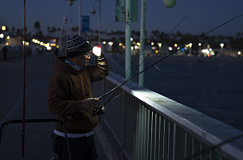 A person wearing a brown hoodie and knit hat is night fishing on a pier, illuminated by a headlamp. They are holding a fishing rod and standing by a railing, with the ocean and city lights visible in the background.