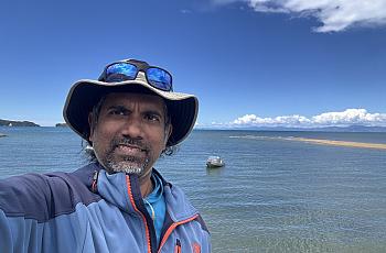 An Sri Lankan Tamil man stands before an expanse of water smiling in a sunhat