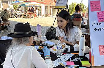 A woman speaks with another person at an outdoor table with pamphlets and notes, next to a mental health sign, at a sunny community market.