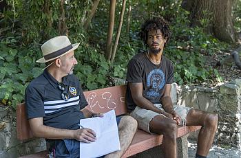 A young man sits on a park bench speaking with an older man who holds a notebook and pen. They appear to be engaged in a serious conversation surrounded by greenery.