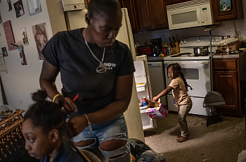 Person doing a child's hair while another child pulls a box out of the refrigerator