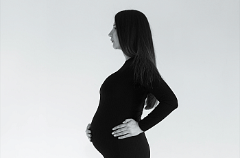 A young Asian Indian woman with long dark hair stands in profile, pregnant, in a tight black dress