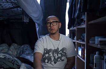 A young Korean man with black glasses sits on his bed